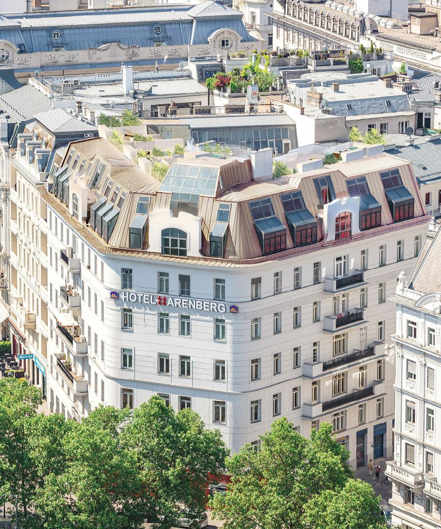 Luftaufnahme eines weißen Stadthotels mit einem geschwungenen Zinkdach und den umliegenden städtischen Gebäuden.
